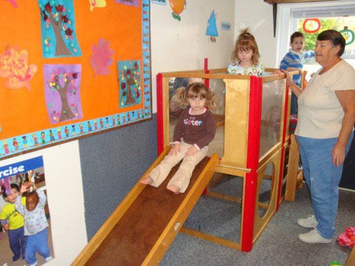Child on indoor playground slide with adults nearby in a classroom setting