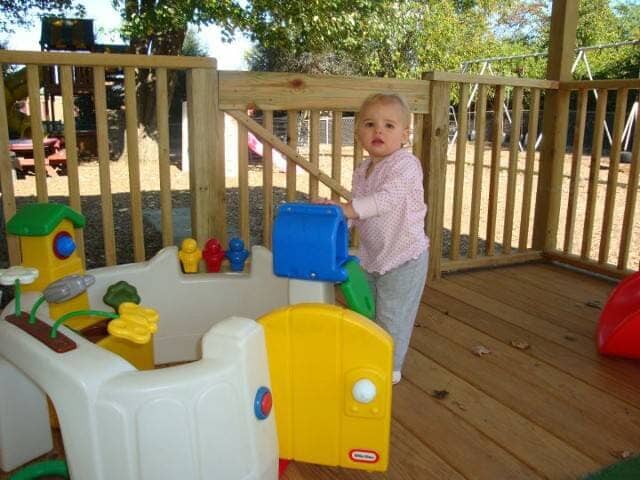 Toddler standing on a wooden deck beside a colorful plastic play kitchen outdoors