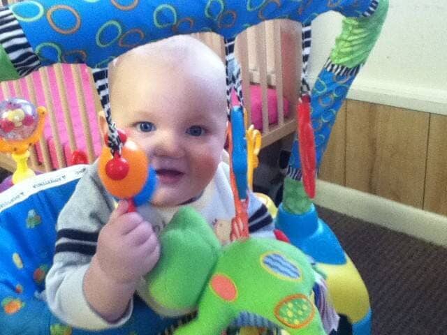 Smiling baby in a colorful activity center, holding a toy rattle.