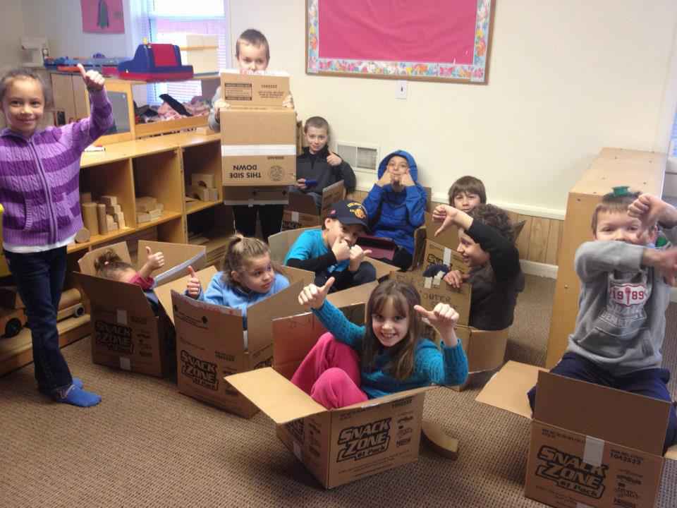 Children playing in cardboard boxes in a classroom, some smiling and waving.