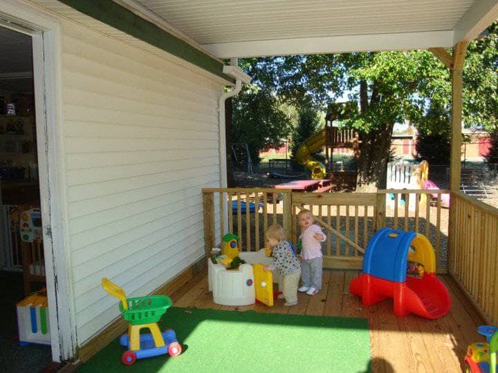 Covered porch with children’s toys, a railing, and a sunny yard beyond