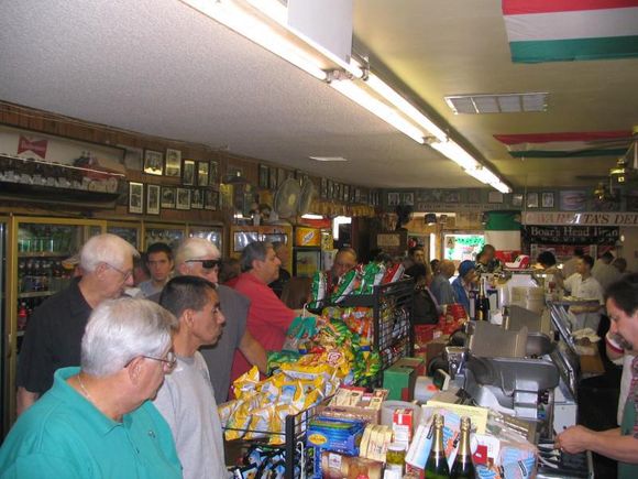 Busy deli/grocery checkout line with shoppers and colorful snack displays inside a small store