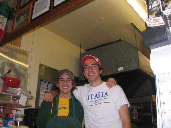 Two smiling people stand together in a small indoor shop, one in a green jacket and one in a white “ITALIA” shirt.