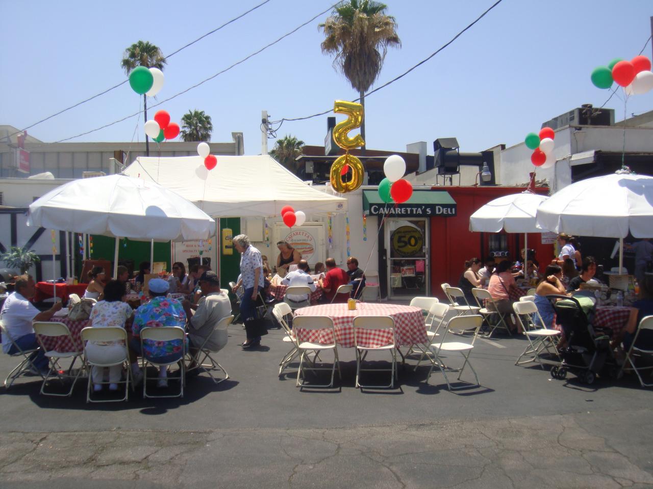 Outdoor restaurant patio with diners under white umbrellas and red-and-green balloons on a sunny day