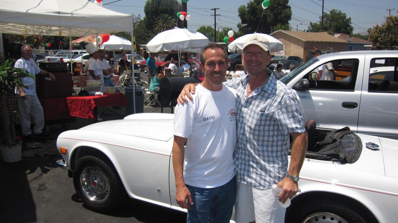 Two men posing beside a white classic convertible at an outdoor car show.