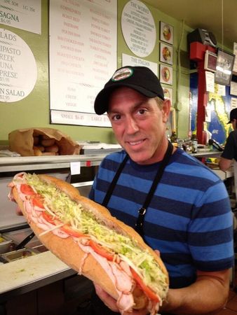 Man in striped shirt and cap holding a giant sub sandwich inside a deli counter