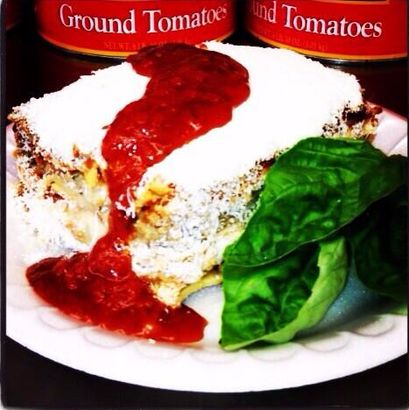 Slice of lasagna on a plate, topped with white cheese and red tomato sauce, with basil leaves beside it.
