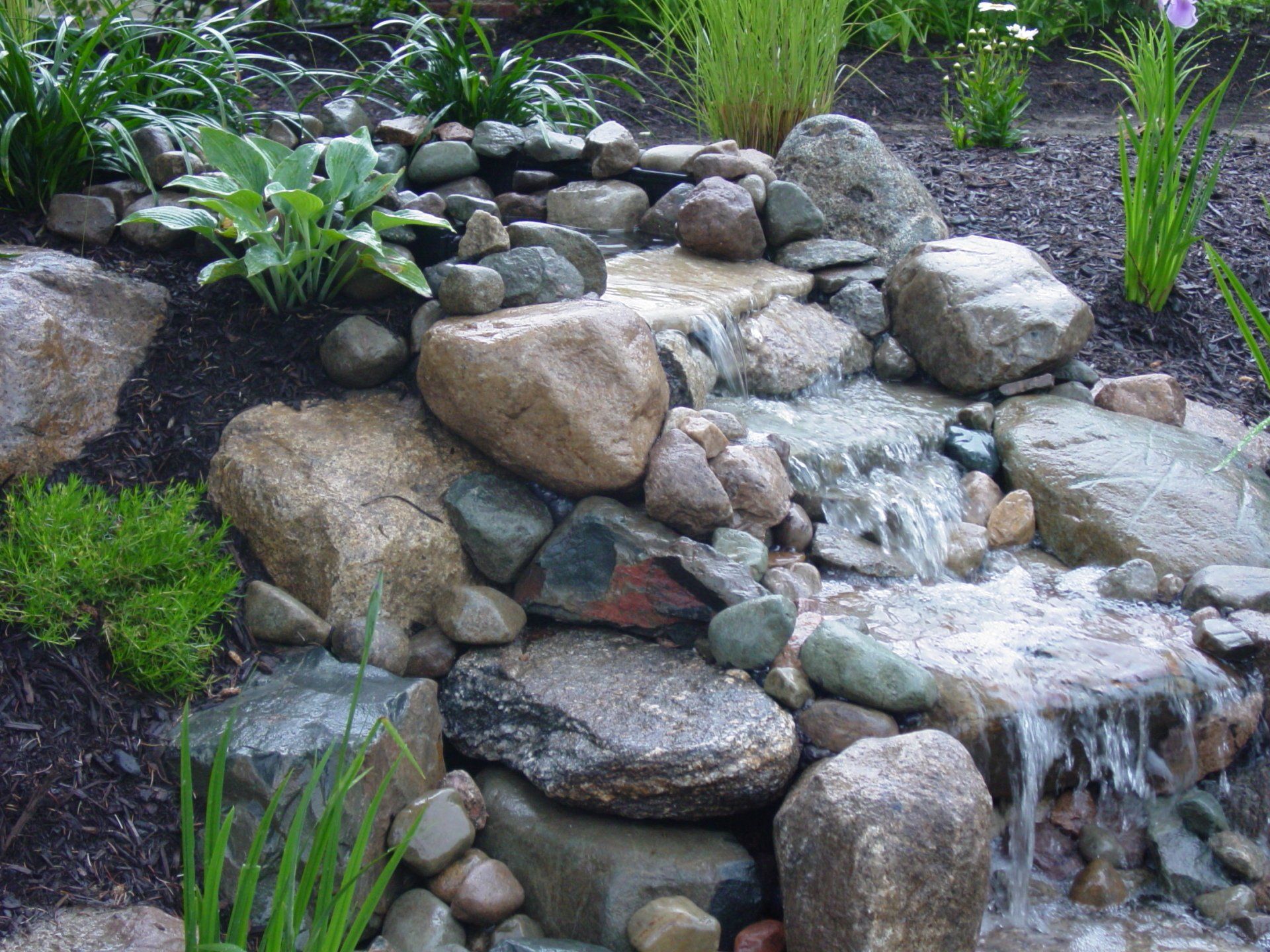 A small waterfall is surrounded by rocks and plants in a garden.