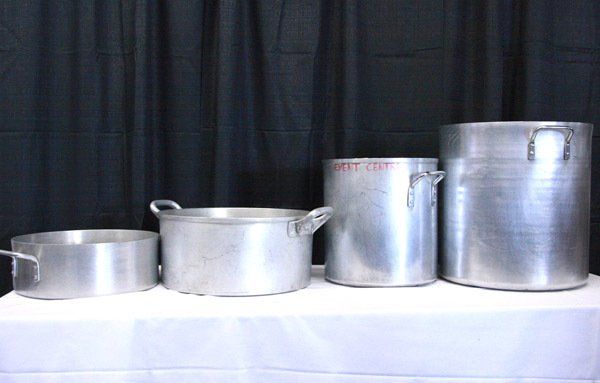 Four aluminum stock pots and containers lined up on a white table before a black curtain