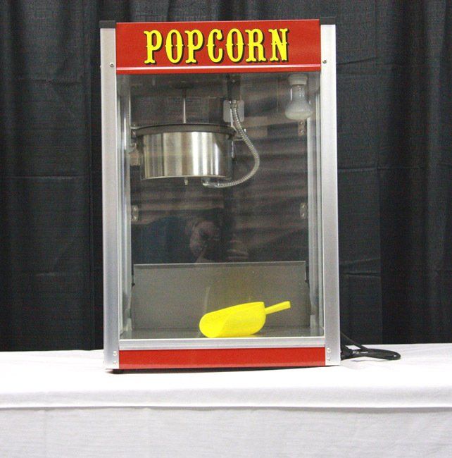 Popcorn machine with red top sign, clear glass front, and yellow scoop inside on a table