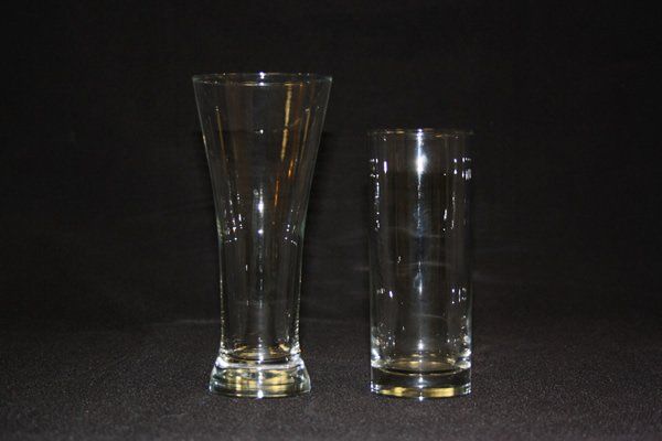 Two clear drinking glasses on a dark background, one tall and flared, one short and cylindrical.