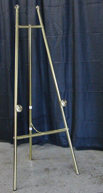 Tall brass easel with adjustable legs and white knobs against a dark curtain backdrop