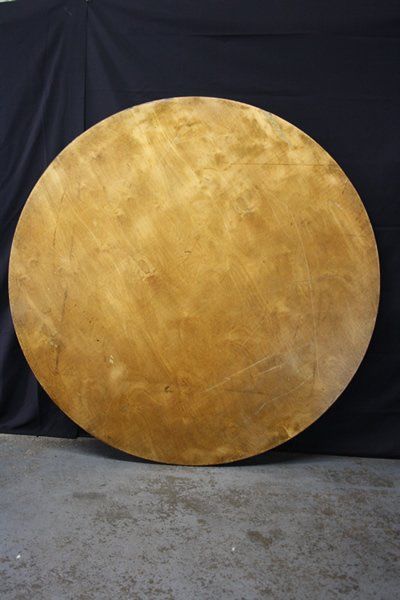 Large round golden-brown wooden disc leaning against a dark backdrop on a concrete floor