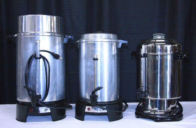 Three stainless steel beverage urns on black stands against a dark backdrop.