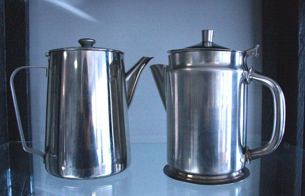 Two stainless steel coffee pots on a reflective surface, one with a long spout and one with a handle and lid