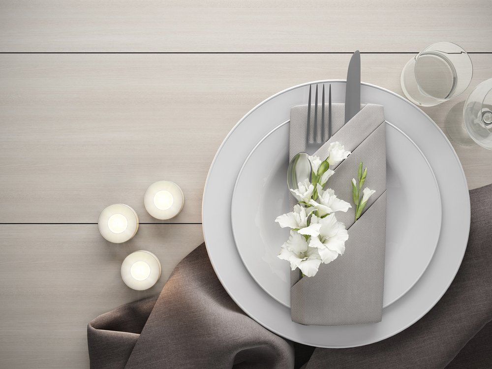 Elegant table setting with white plates, folded gray napkin, flowers, and candles on a light wood table