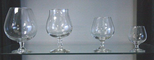 Four empty wine glasses of different sizes lined up on a glass shelf against a gray wall