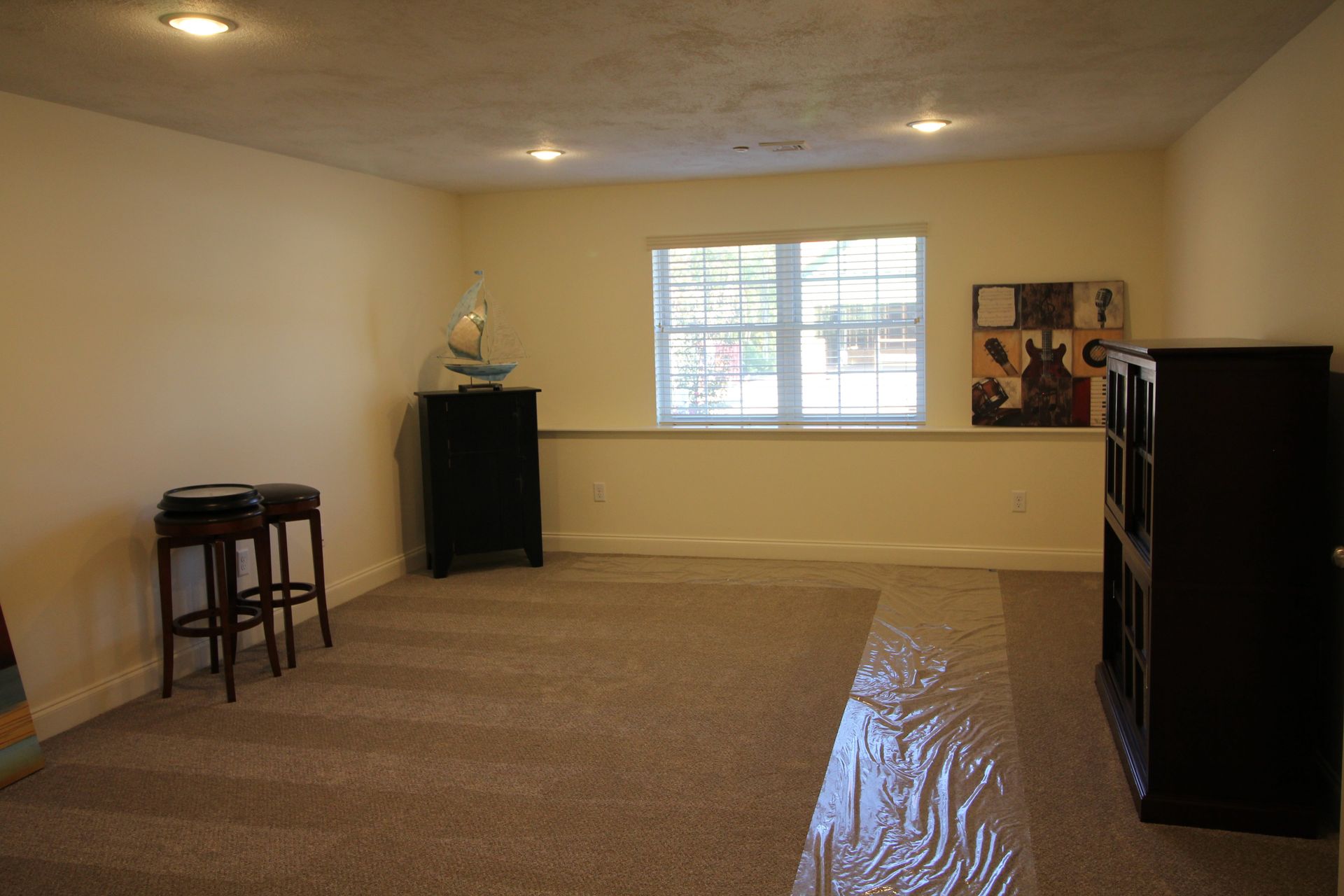 An empty room with a carpeted floor , two stools , a bookshelf and a window.