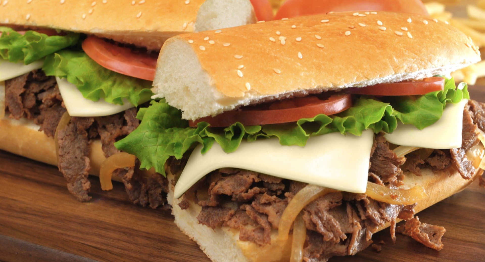 Two sub sandwiches with beef , cheese , lettuce and tomatoes on a wooden cutting board.