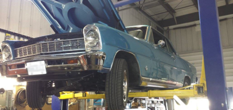 A blue car with the hood up is on a lift in a garage.