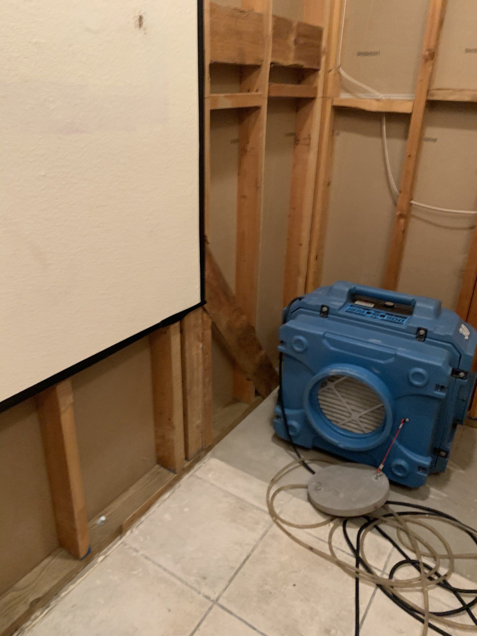 Damaged interior wall with exposed studs, blue fan, and loose cords on a tiled floor.