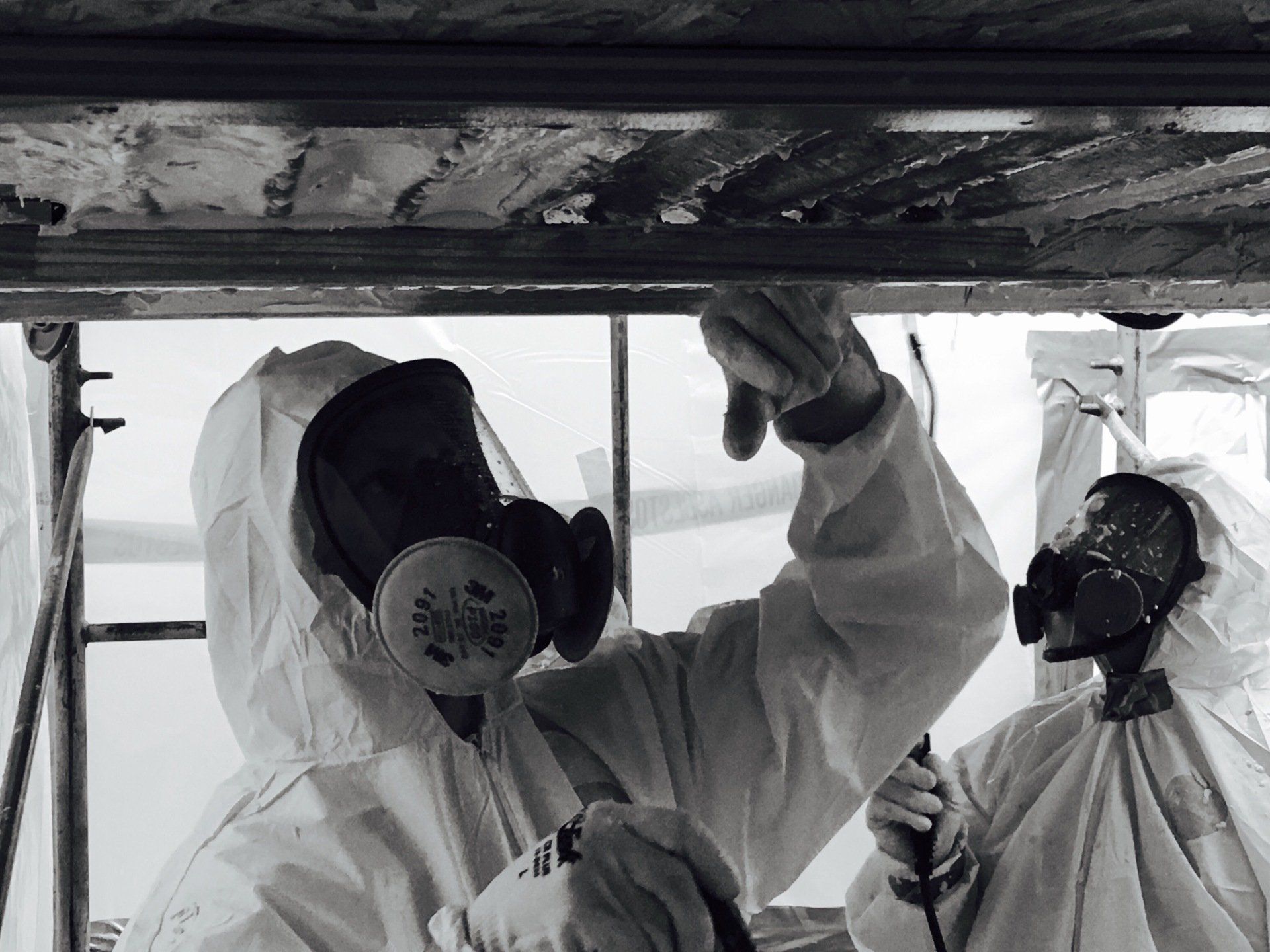 Two workers in protective suits spraying chemicals on a ceiling beam in an industrial setting
