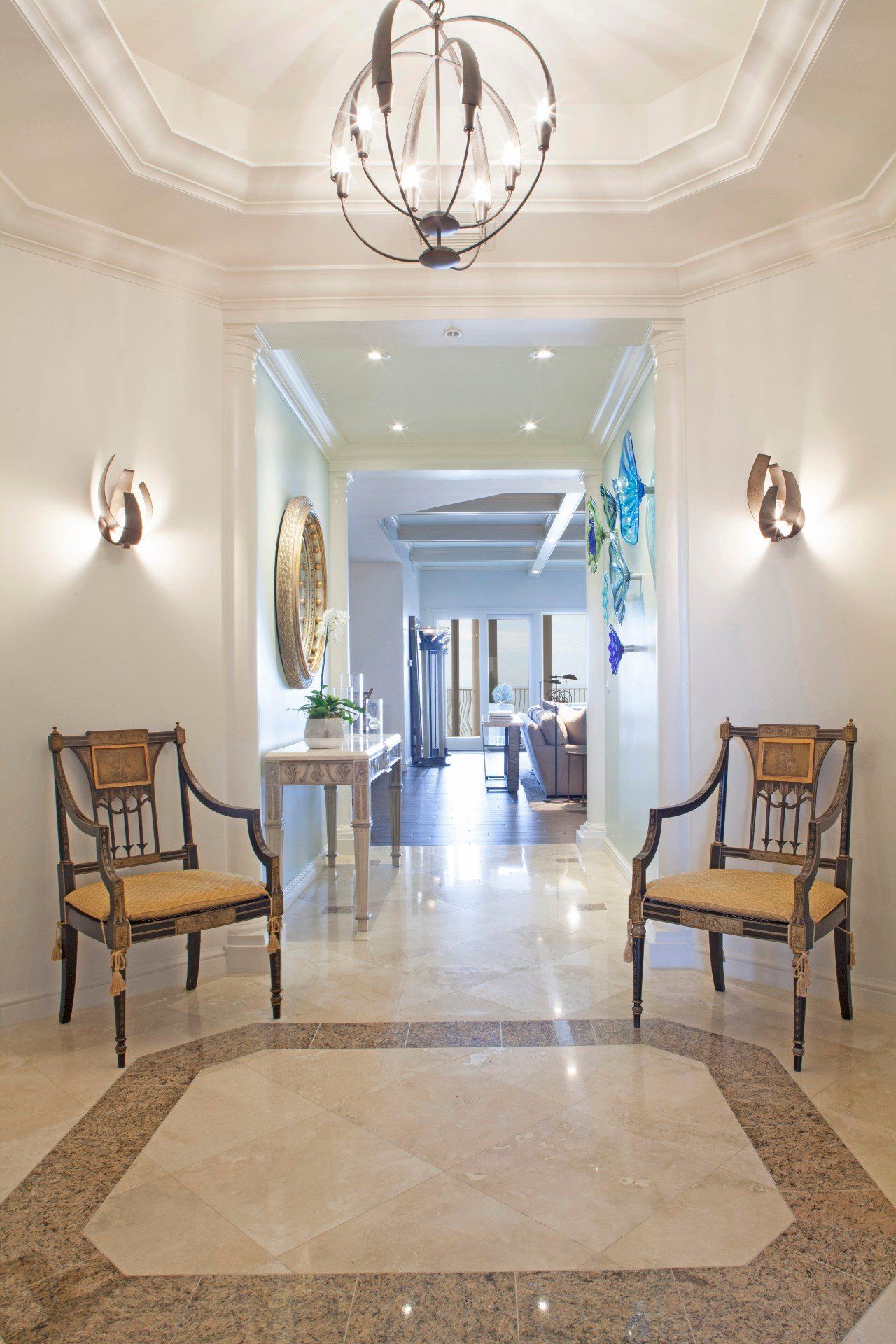 Elegant hotel foyer with marble floor, upholstered chairs, wall sconces, and a chandelier leading to a bright hallway