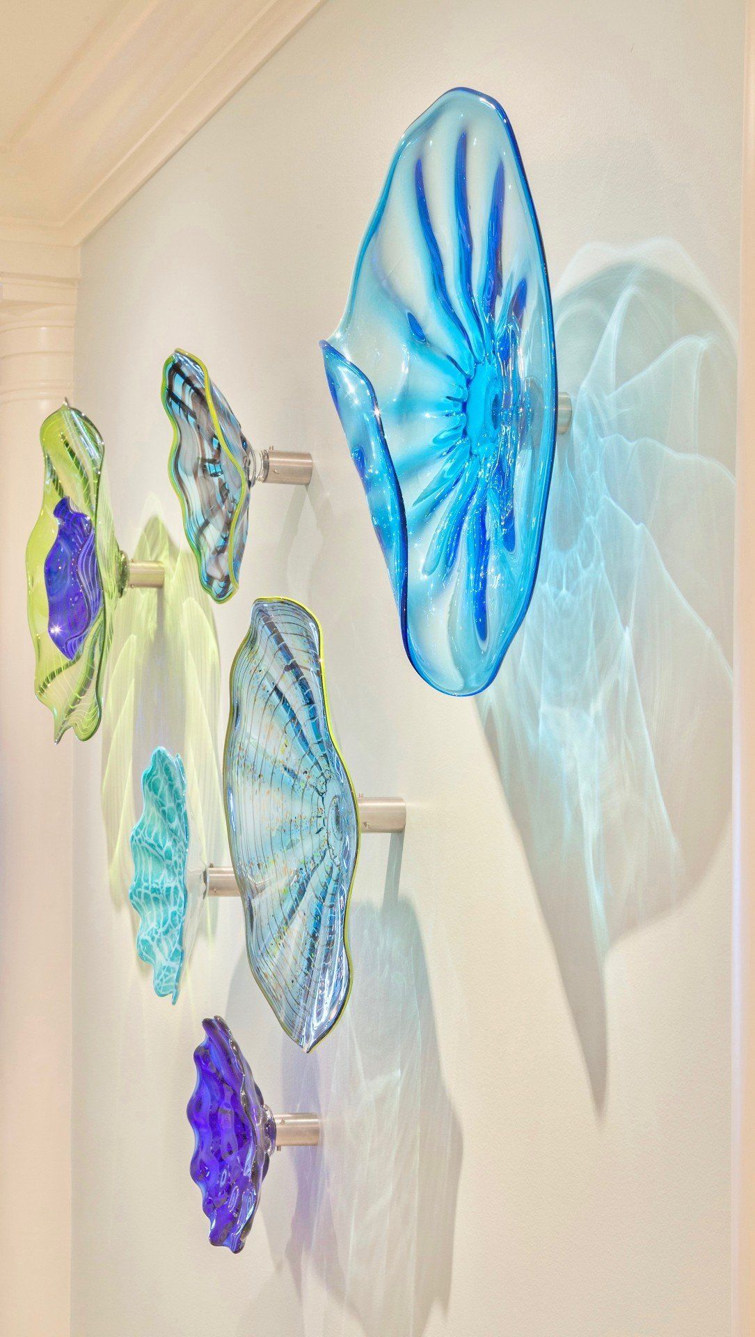 Blue and green glass wall art with layered butterfly and flower-like shapes on a cream wall