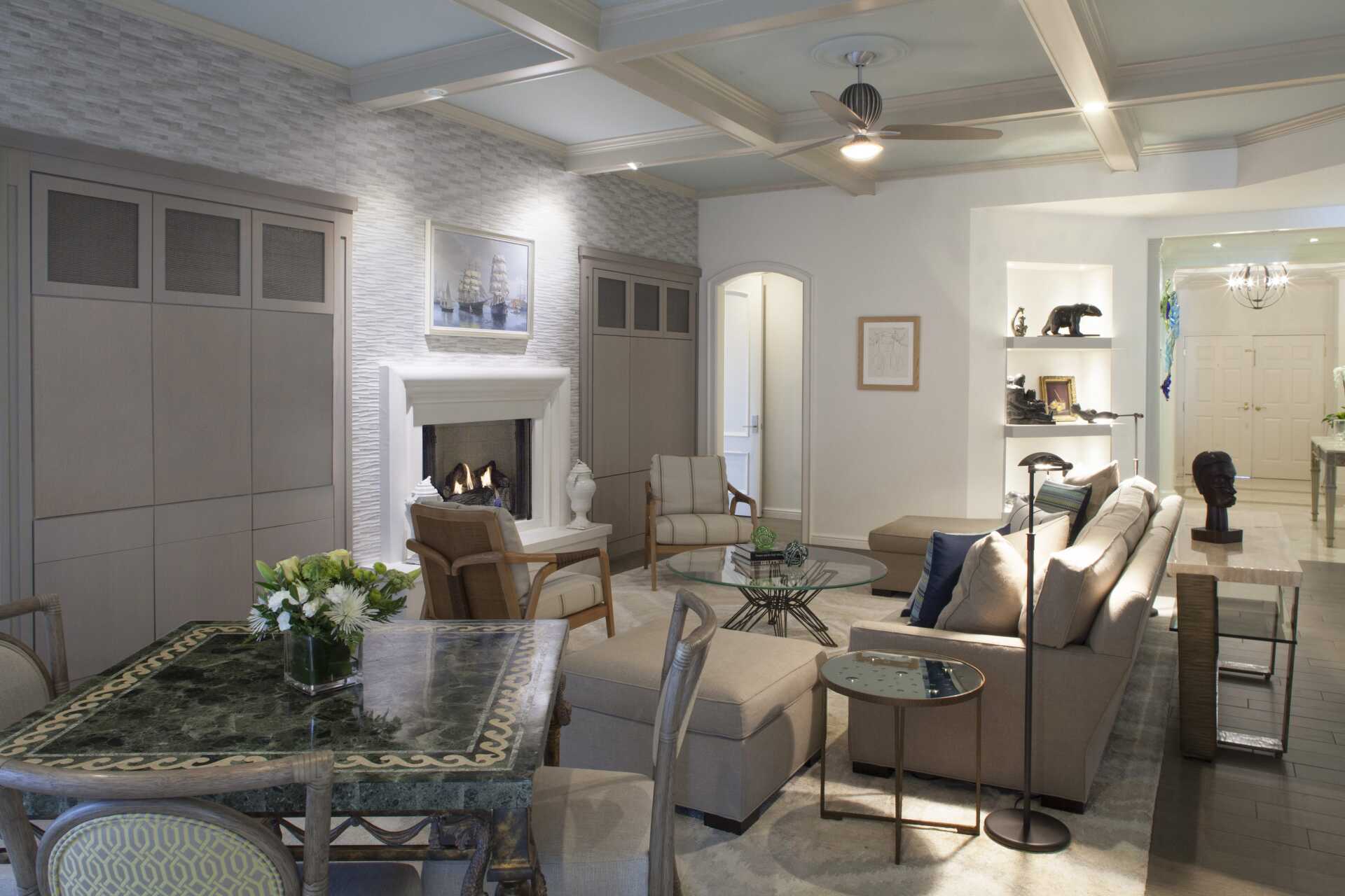 Bright living room with beige sofas, glass coffee table, fireplace, and built-in shelves, lit by soft overhead light