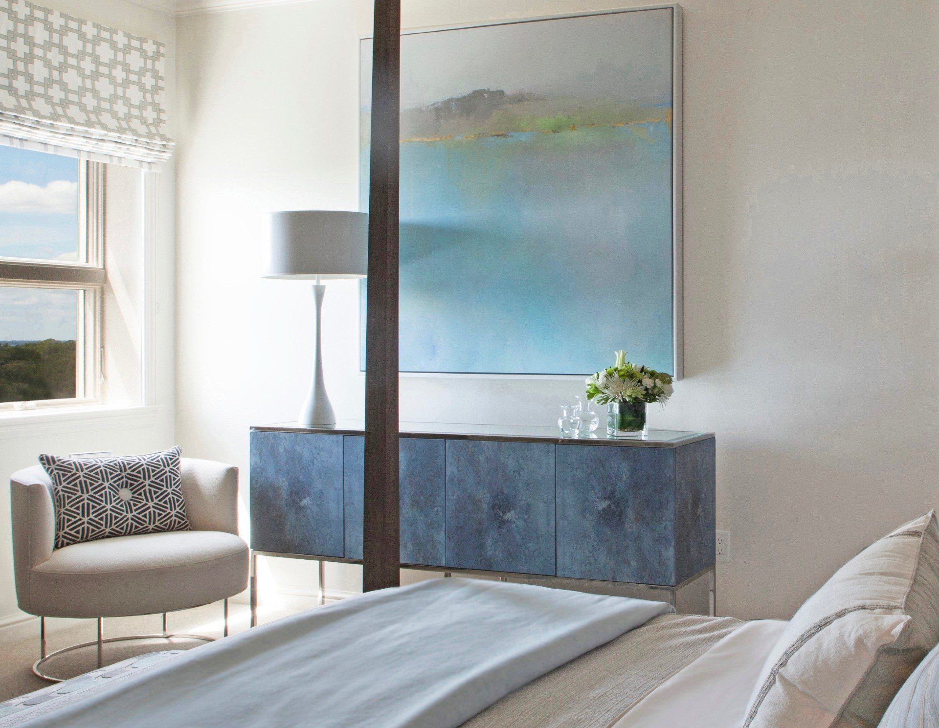 Modern bedroom with blue dresser, large abstract art, armchair, window, and bed in the foreground