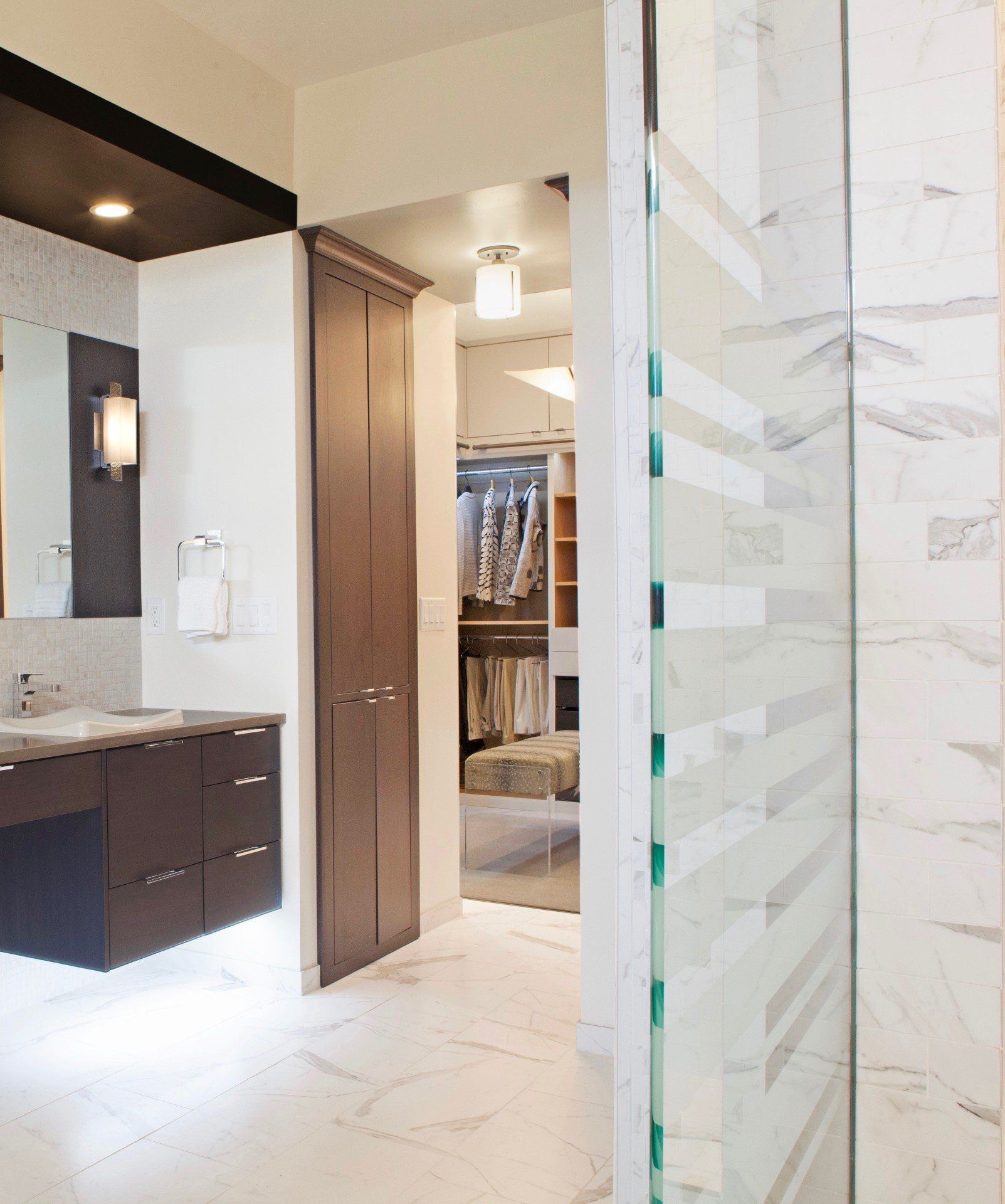 Modern bathroom with dark vanity, white marble tile, and open walk-in closet visible through the doorway