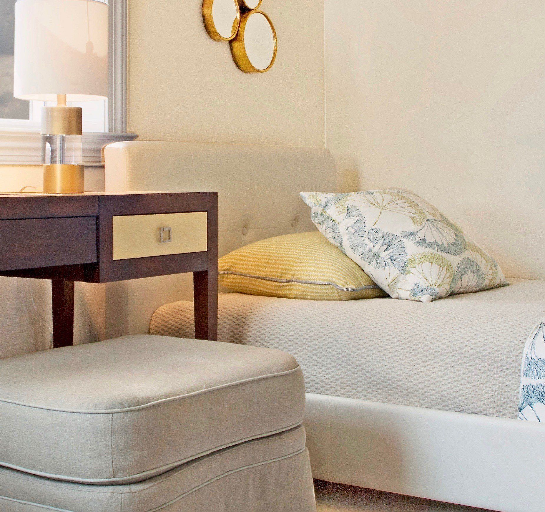 Cozy bedroom corner with a bed, bedside table lamp, patterned pillows, and a beige ottoman.