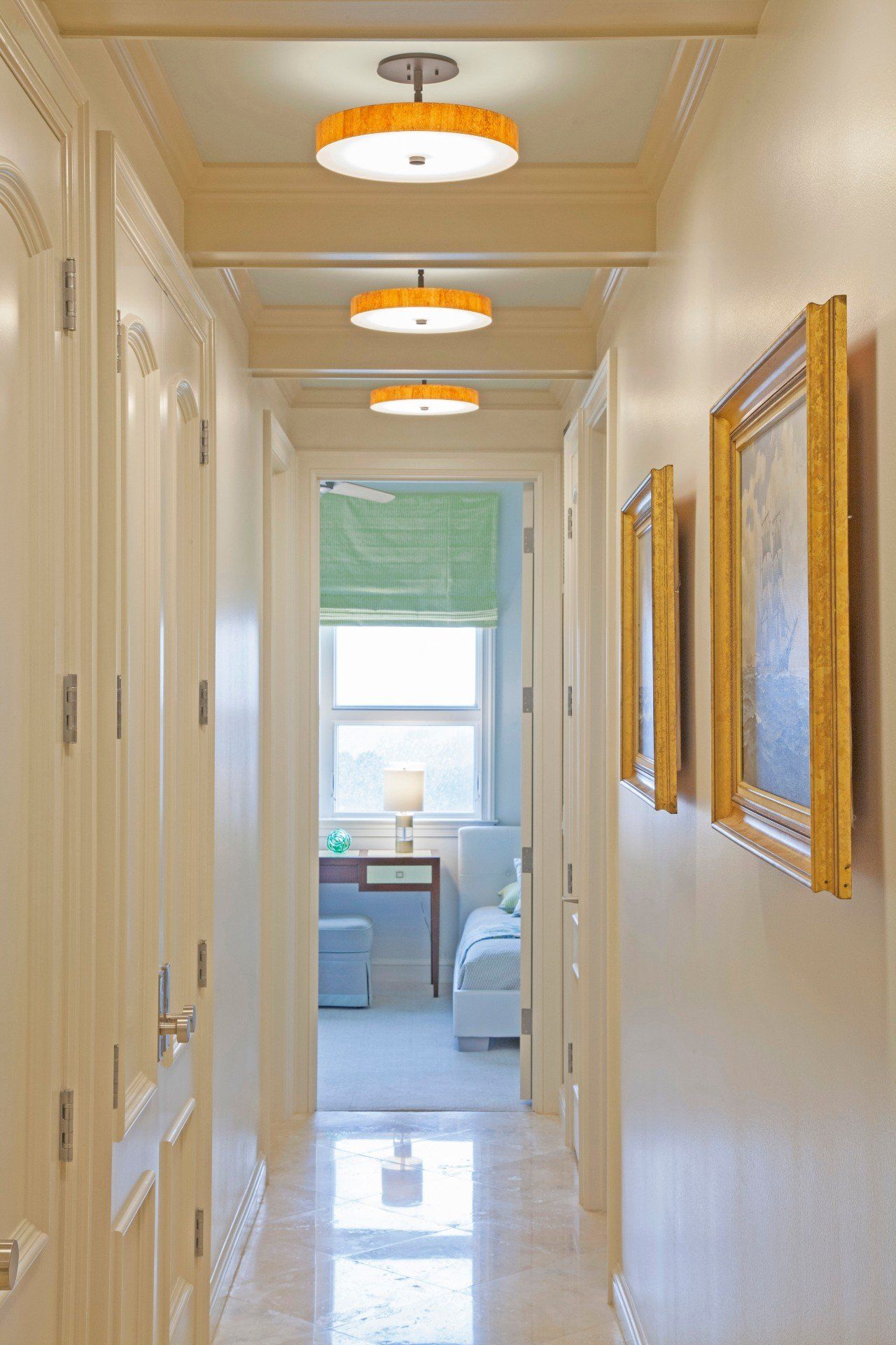 Narrow cream hallway with framed gold mirrors, ceiling lights, and a bright window at the far end