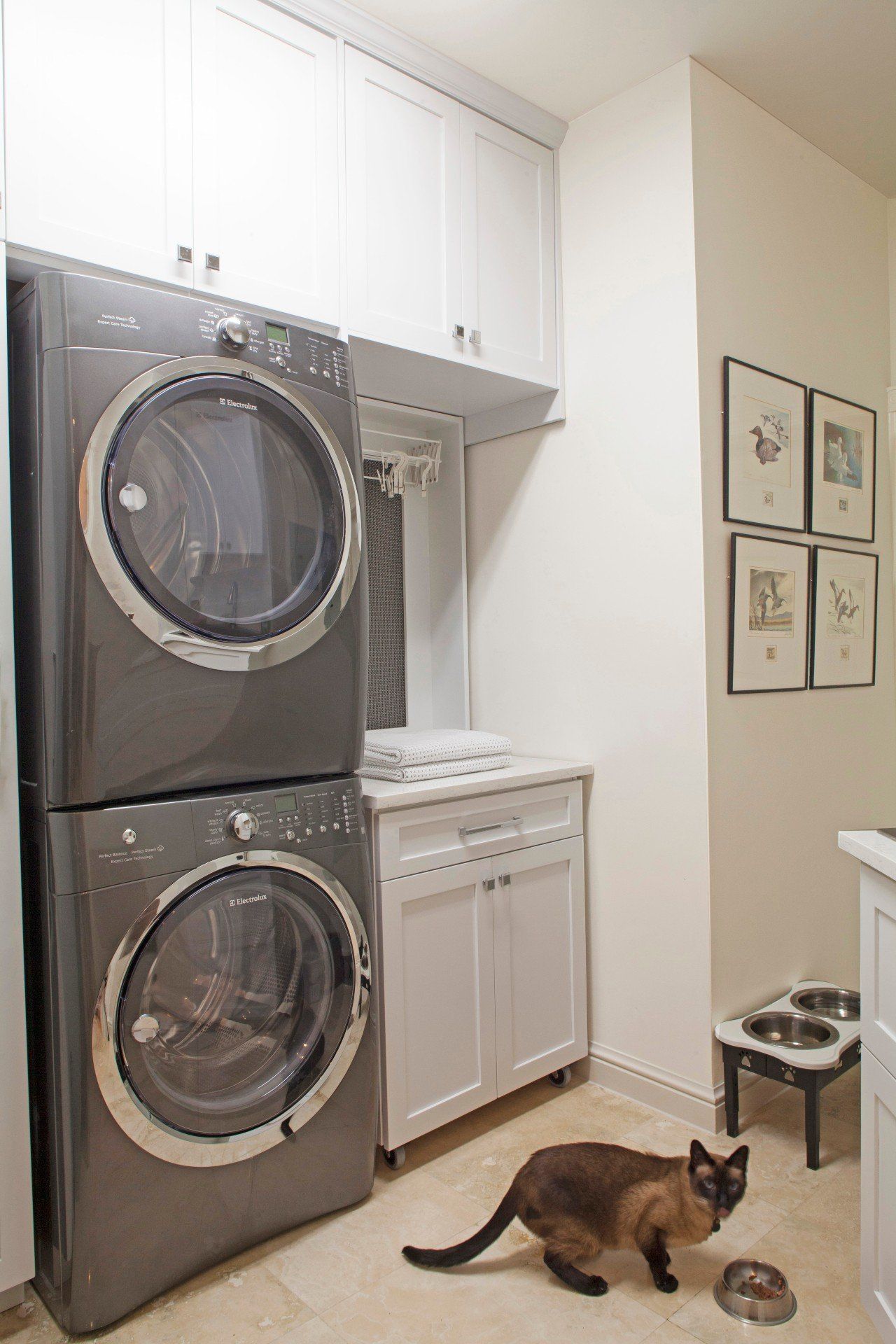 Laundry room with stacked washer and dryer; a cat sits on the floor near a small stool.