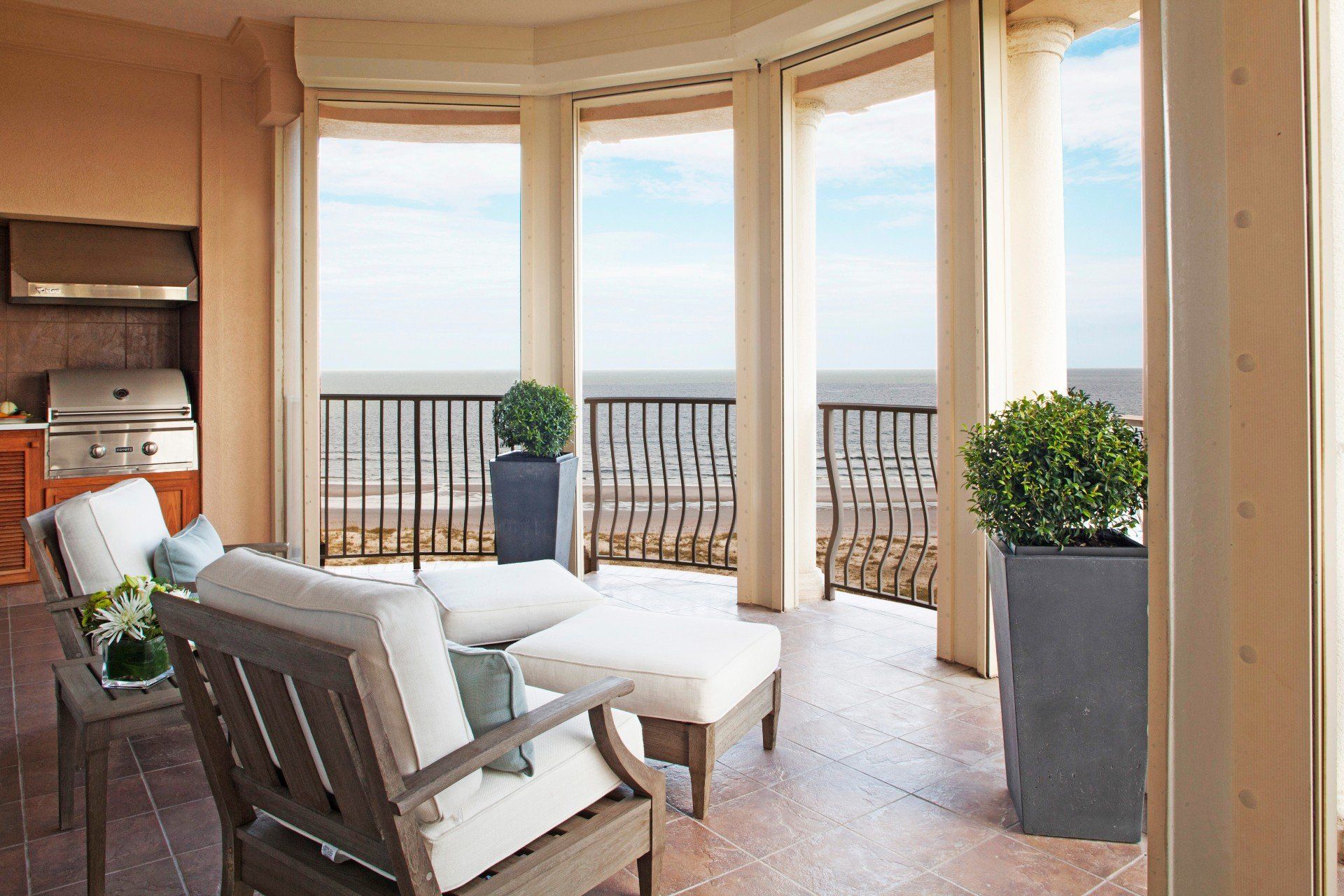 Balcony lounge with cushioned chairs, white table, potted plants, and ocean view through tall columns