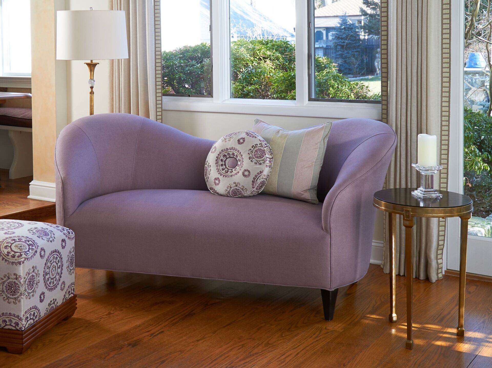 Lavender loveseat in a bright living room with patterned pillows, lamp, and wooden floor
