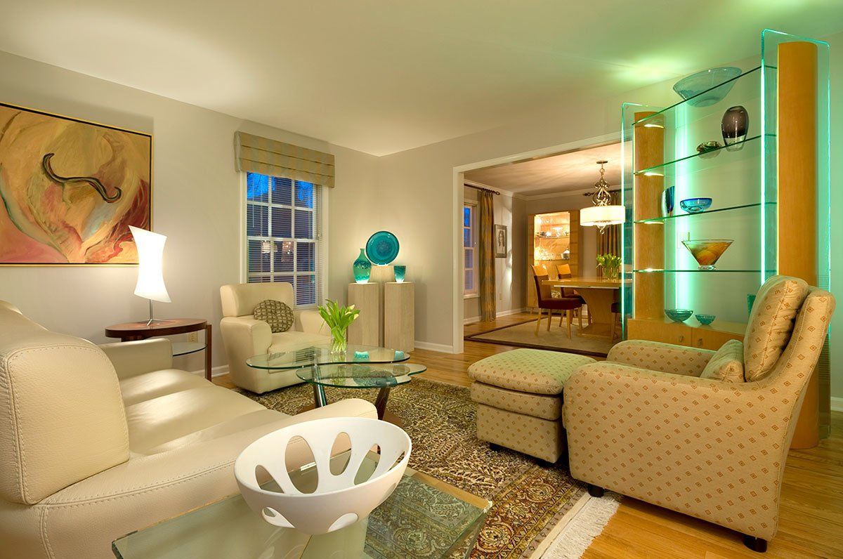 Stylish living room with beige seating, glass accents, and soft green lighting, viewed into a dining area