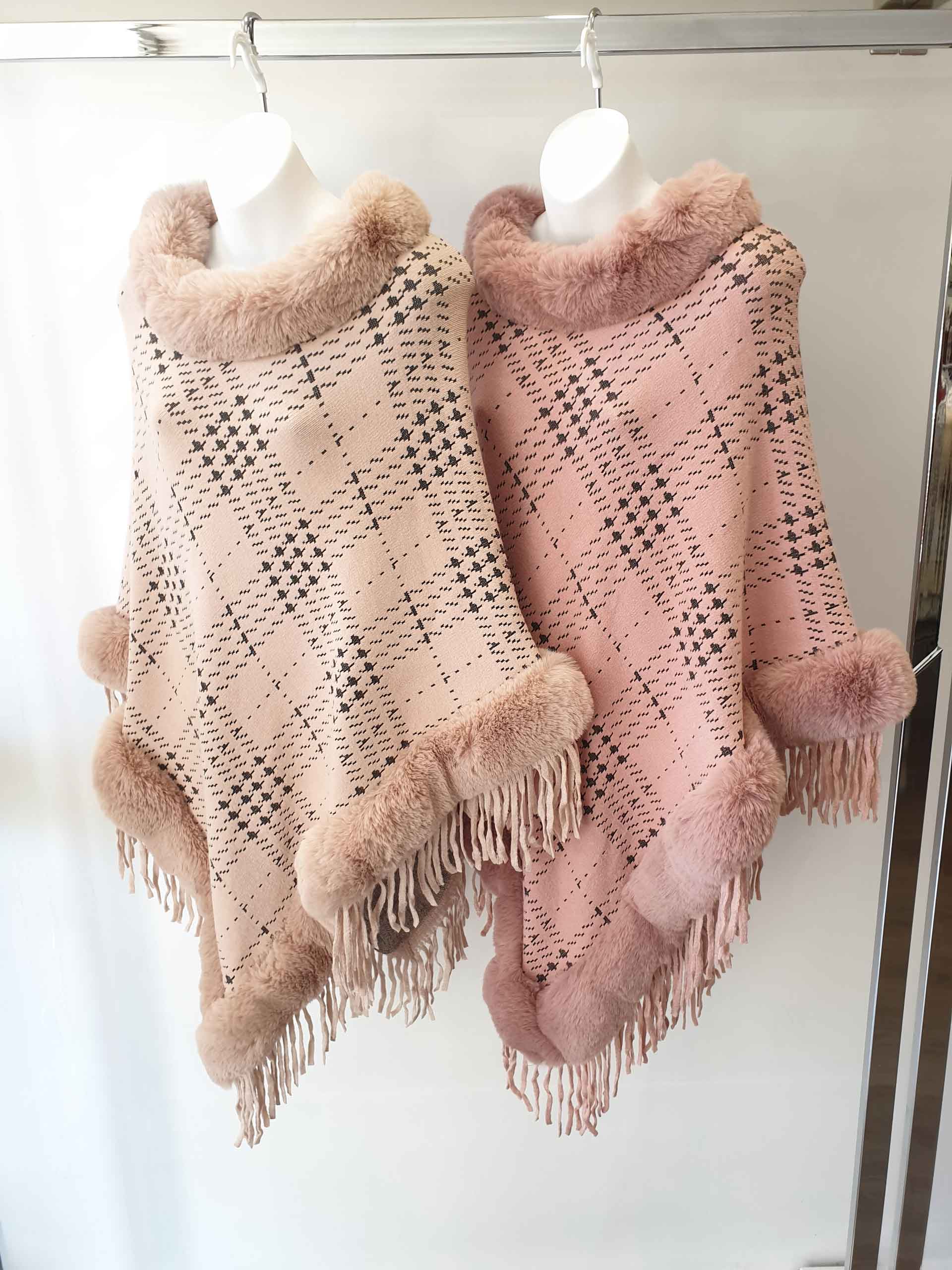 Two pink and beige ponchos with fur trim and fringe hanging on a rack.