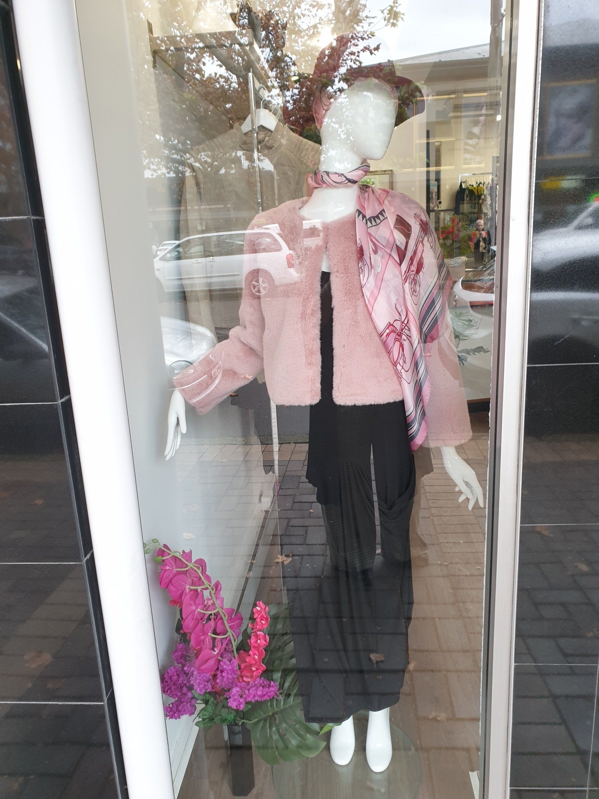 Mannequin in shop window, wearing pink jacket, scarf, black pants, and hat. Flowers in front.