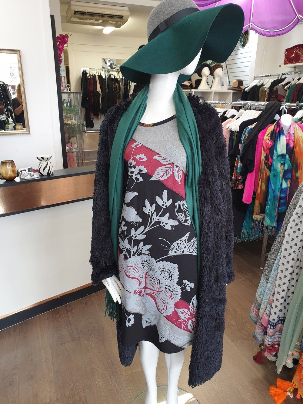 Mannequin in a shop window wearing a floral dress, green scarf, dark coat, and a wide-brimmed hat.