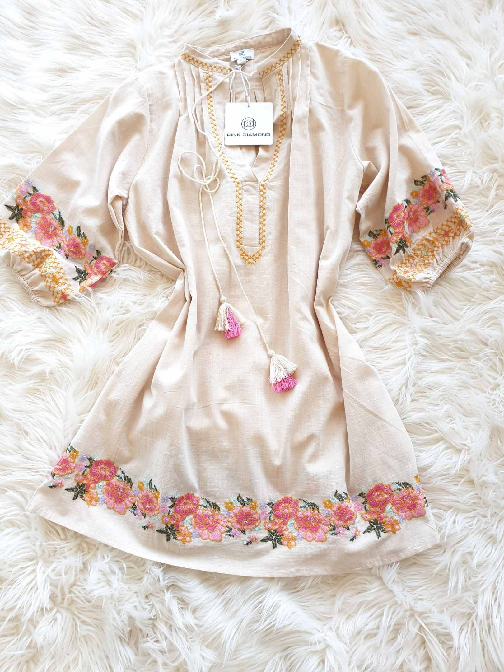 Beige tunic dress with floral embroidery at the hem and sleeves, tassels, and a gold necklace.