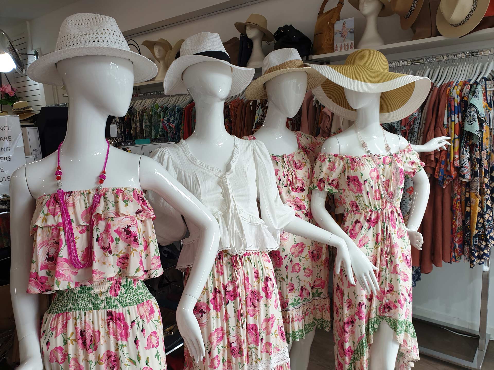 Four mannequins wearing summer outfits and hats in a clothing store, with floral dresses and white tops.