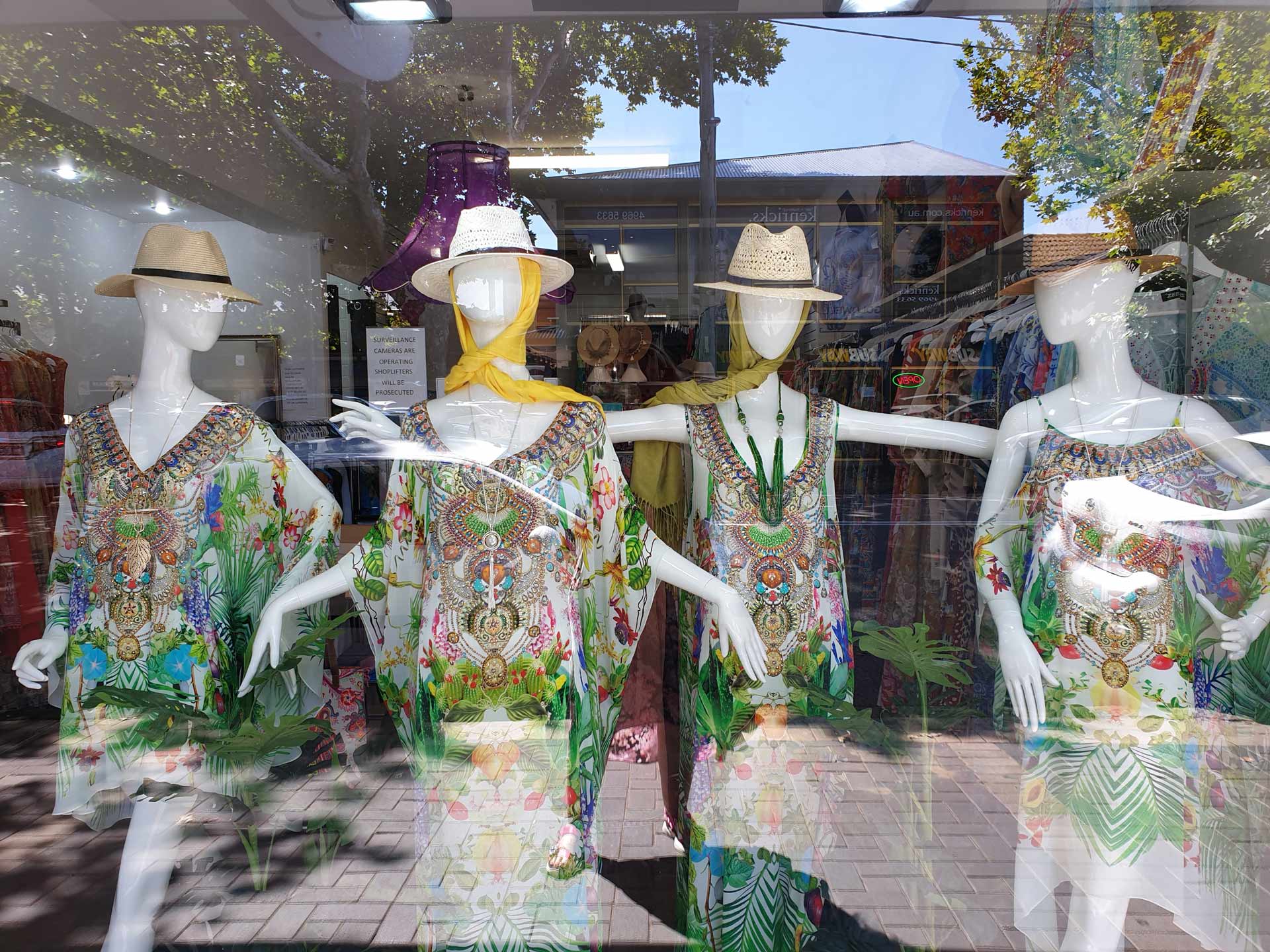Four mannequins in a shop window display wearing colorful patterned dresses and straw hats.