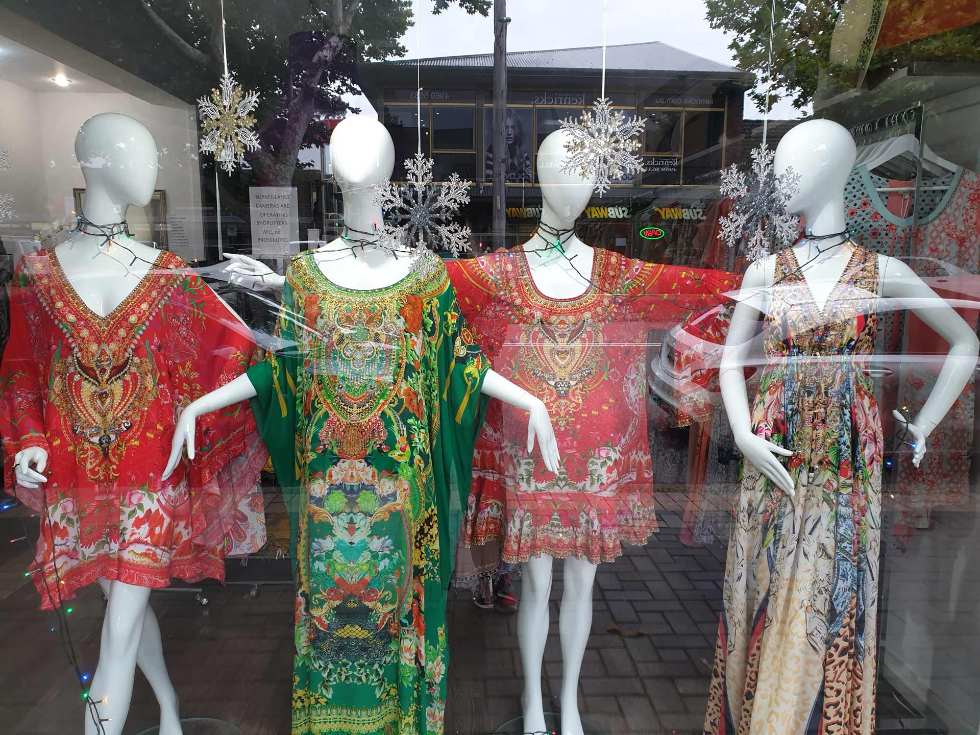 Four mannequins in a store window, wearing colorful patterned dresses. Festive snowflakes hang.