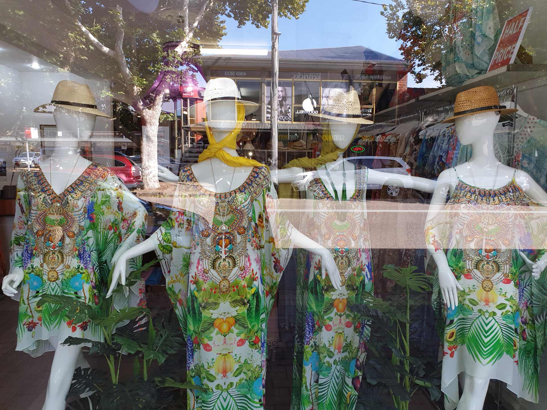 Mannequins wearing patterned tunics and hats in a shop window, with street and other shops reflected in the glass.