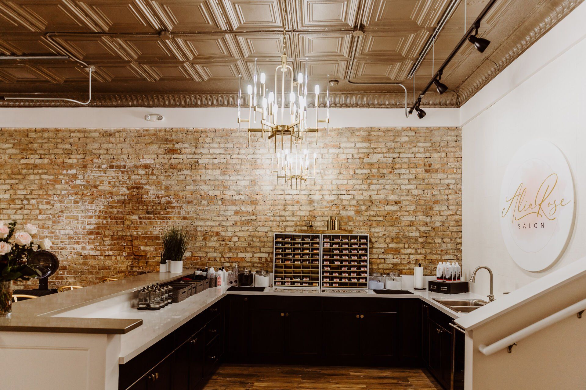 A beauty salon interior with exposed brick wall, black cabinets, and a chandelier.