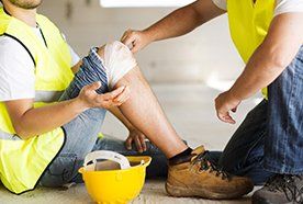 A builder sitting next to his yellow hard hat, with someone putting a bandage on his knee