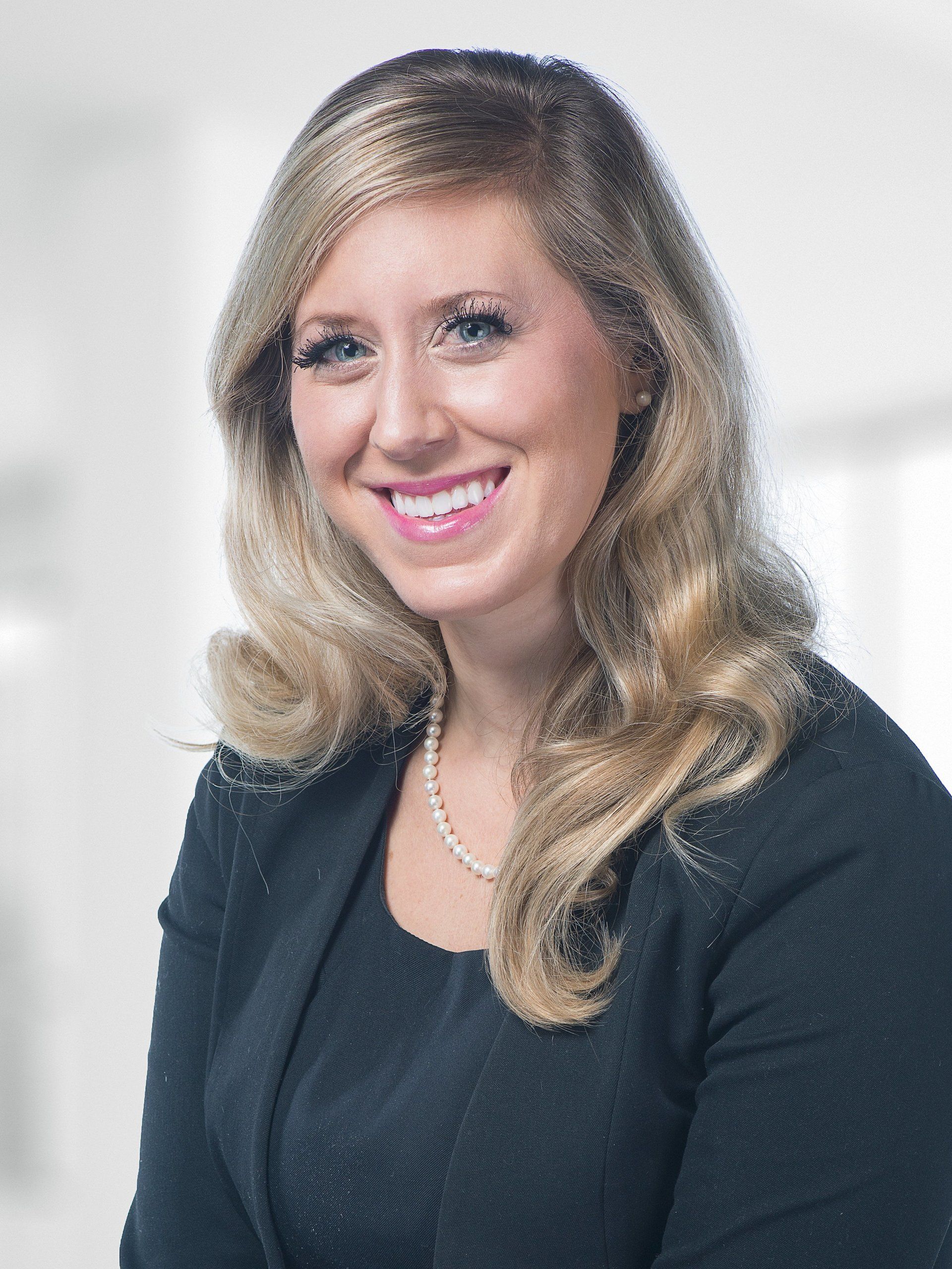 A woman wearing a black jacket and a pearl necklace is smiling for the camera.