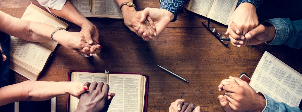 A group of people are sitting around a table with their hands folded in prayer.