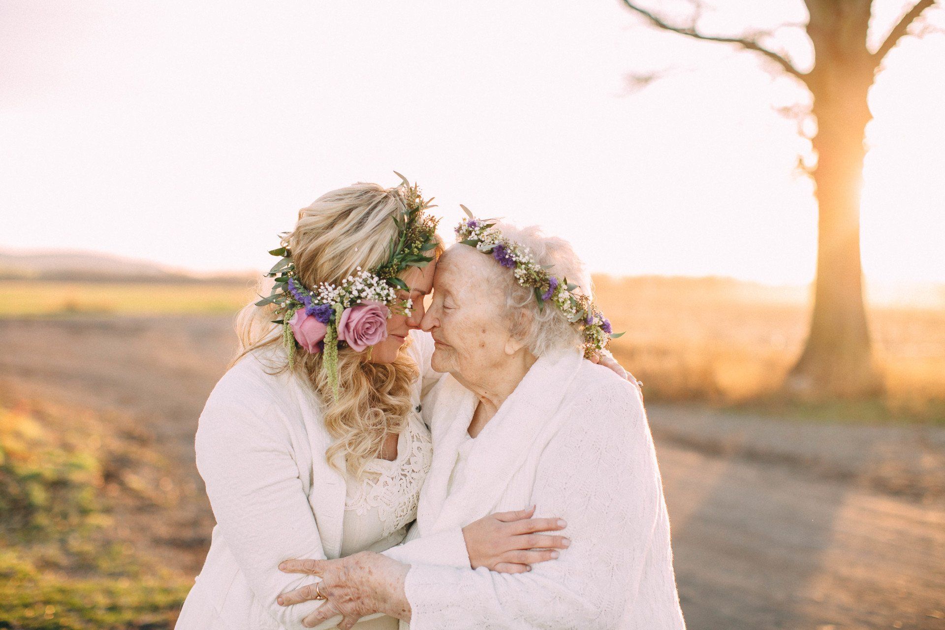 Woman with floral crown embraces older woman, both in white, outdoors at sunset.